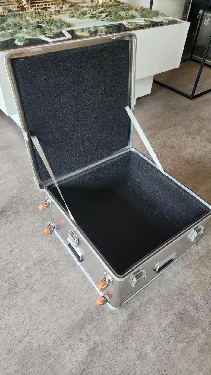 Custom Aluminium Cases Open aluminum flight case with silver metal frame, black foam-padded interior, orange caster wheels, open lid supported by strap, displayed on gray floor in architectural model studio with large building scale models in background