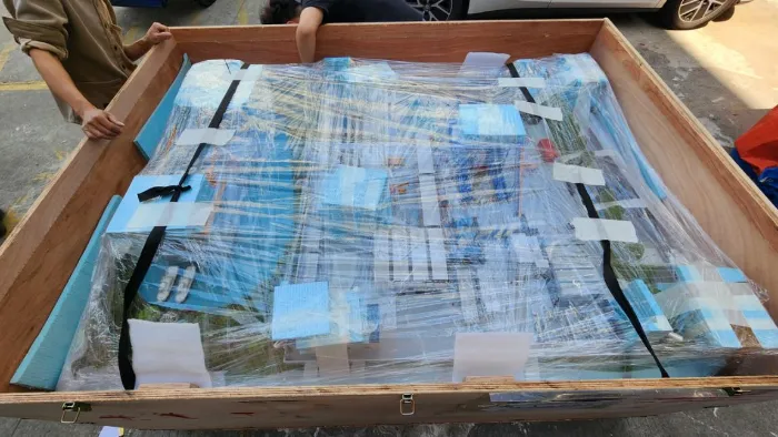 Custom packaging Large wooden export shipping crate held by workers, containing a heavily shrink-wrapped architectural model package secured with black ratchet straps, blue foam padding and white tape layers – professional heavy-duty custom packaging for safe transport