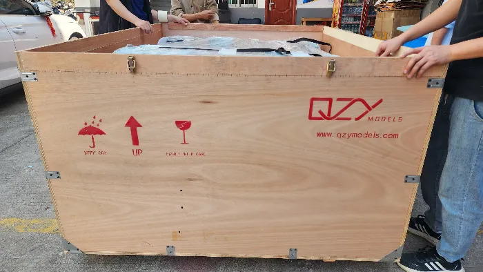 Custom wooden Crate Large plywood export shipping crate with red international handling symbols (fragile, this way up, keep dry), branded QZY MODELS and www.qzymodels.com, held by workers on outdoor pavement – professional heavy-duty wooden packaging