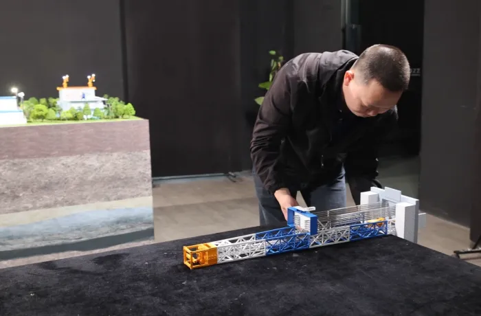 Detachable drilling rig Man assembling detachable drilling rig scale model on black display table, showing modular blue-white-orange metal truss structure with platform sections – professional industrial engineering model demonstration