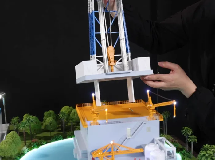 Easy Lighting Connection Hands demonstrating easy plug-in lighting connection on detailed offshore drilling rig scale model: attaching illuminated blue-white tower section with glowing LED lights onto yellow-crane platform base – modular lighted industrial model assembly
