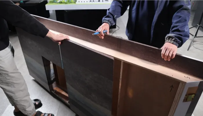 Foldable base structure Two workers assembling large foldable plywood display base with dark wood-grain finish, one marking precise alignment with blue marker while holding edges – modular platform with cutouts and orientation arrow for architectural and industrial scale models