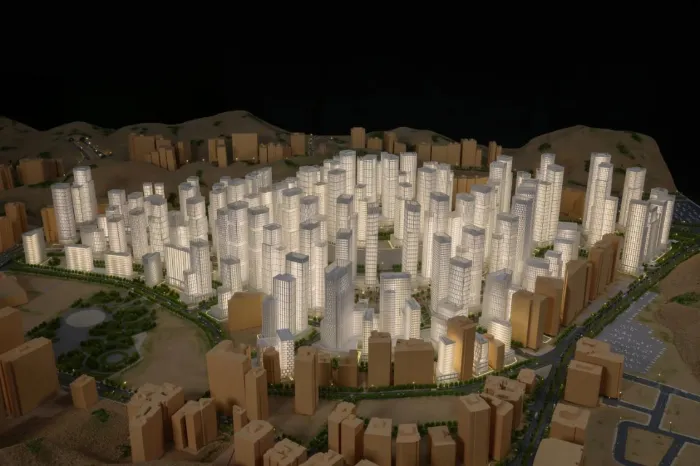 Maqueta a escala de una ciudad moderna, iluminada y con gran nivel de detalle, que presenta un denso conjunto de rascacielos blancos brillantes rodeados de edificios bajos de color beige, sobre un terreno desértico montañoso realista con parques verdes, carreteras, un lago e infraestructura sinuosa: una exhibición profesional de un plan maestro de desarrollo urbano.