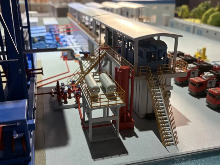 Industrial Processing Plant Model Close-up of highly detailed industrial processing plant scale model with intricate red and blue piping systems, elevated platforms with yellow safety railings, staircases, cylindrical tanks, control buildings and emergency vehicles on display base