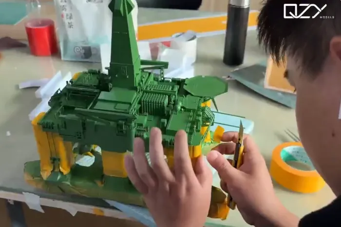 Offshore Platform Model Crafting Craftsman carefully detailing green and yellow offshore platform scale model with precision craft knife on workshop bench, showing intricate deck structures, helipad and support legs – professional model making process by QZY Models