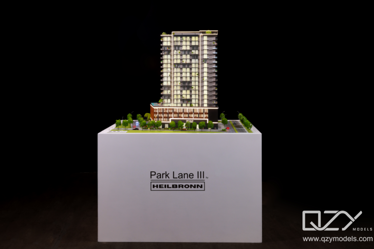  A close-up of an illuminated architectural scale model in a sales suite, with warm directional lighting creating depth and shadow across the building's façade and landscape detail.